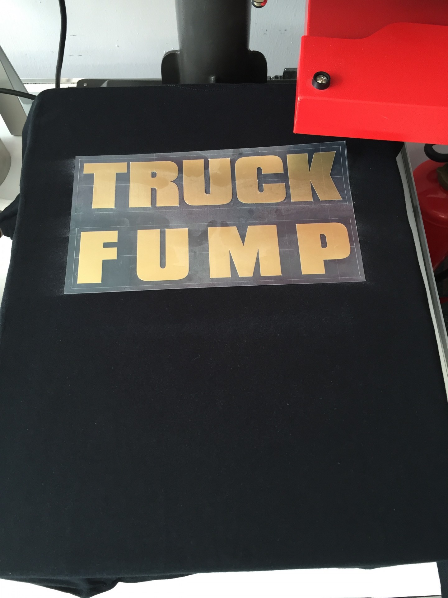 Entgitterter Schriftzug TRUCK FUMP aus goldener Flexfolie auf Trägerfolie, positioniert auf schwarzem T-Shirt in roter Transferpresse