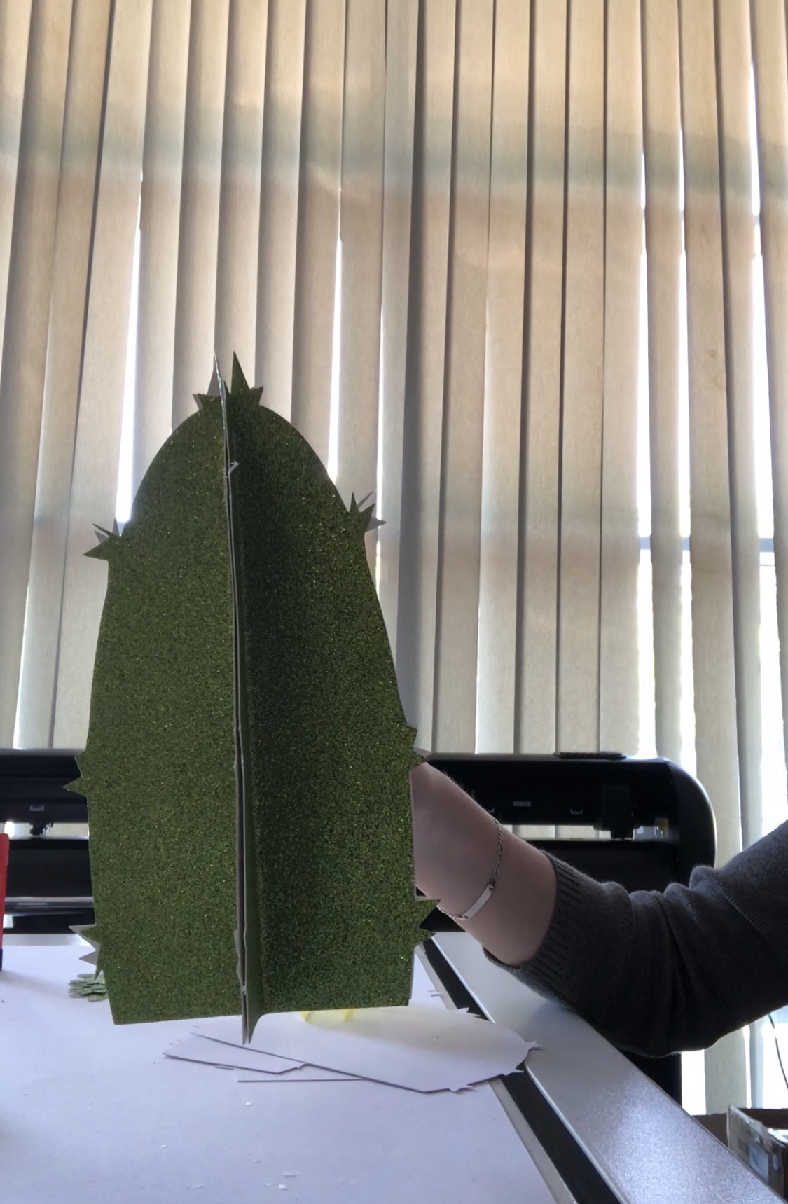 Hand hält zusammengeklebten 3D-Kaktus aus grüner Glitzerpappe mit Stacheln vor einem Schneideplotter, darunter weiße Pappreste