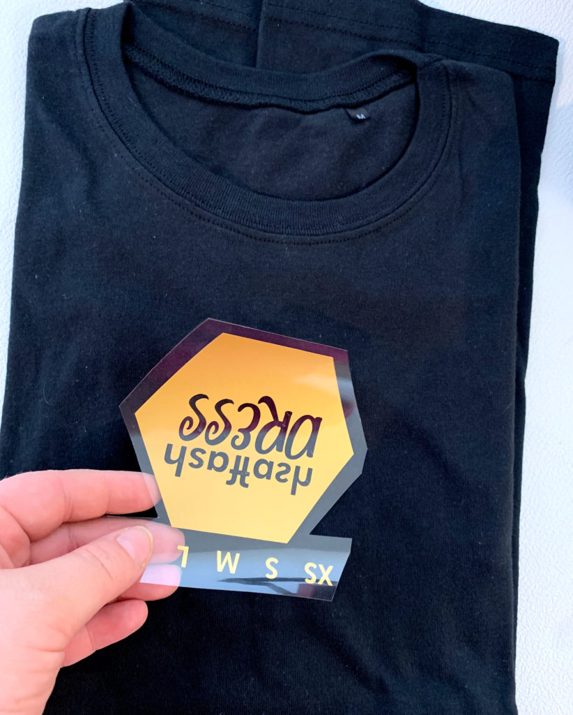 Hand hält gespiegelte Transferfolie mit orangem Sechseck-Motiv und Größen XS bis L über schwarzem T-Shirt, vorbereitet für die TC1