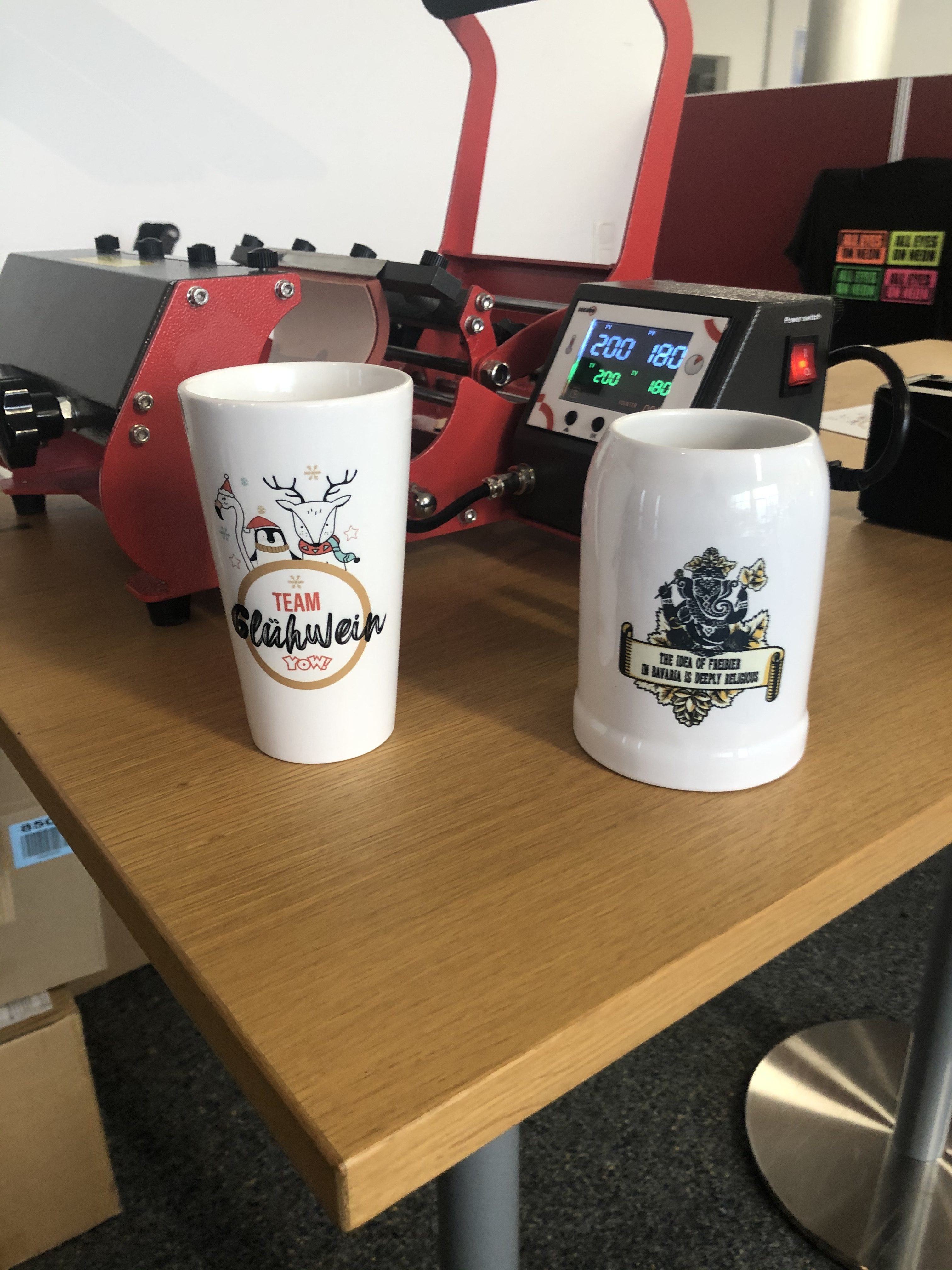 Fertig sublimierter Becher Team Glühwein und Bierkrug mit Ganesha-Motiv vor roter Tassenpresse, Display zeigt 200 °C und 180 s