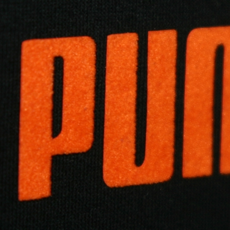 Nahaufnahme orangefarbener Flockfolie mit samtiger Oberfläche als Schriftzug PUMA auf schwarzem Textil