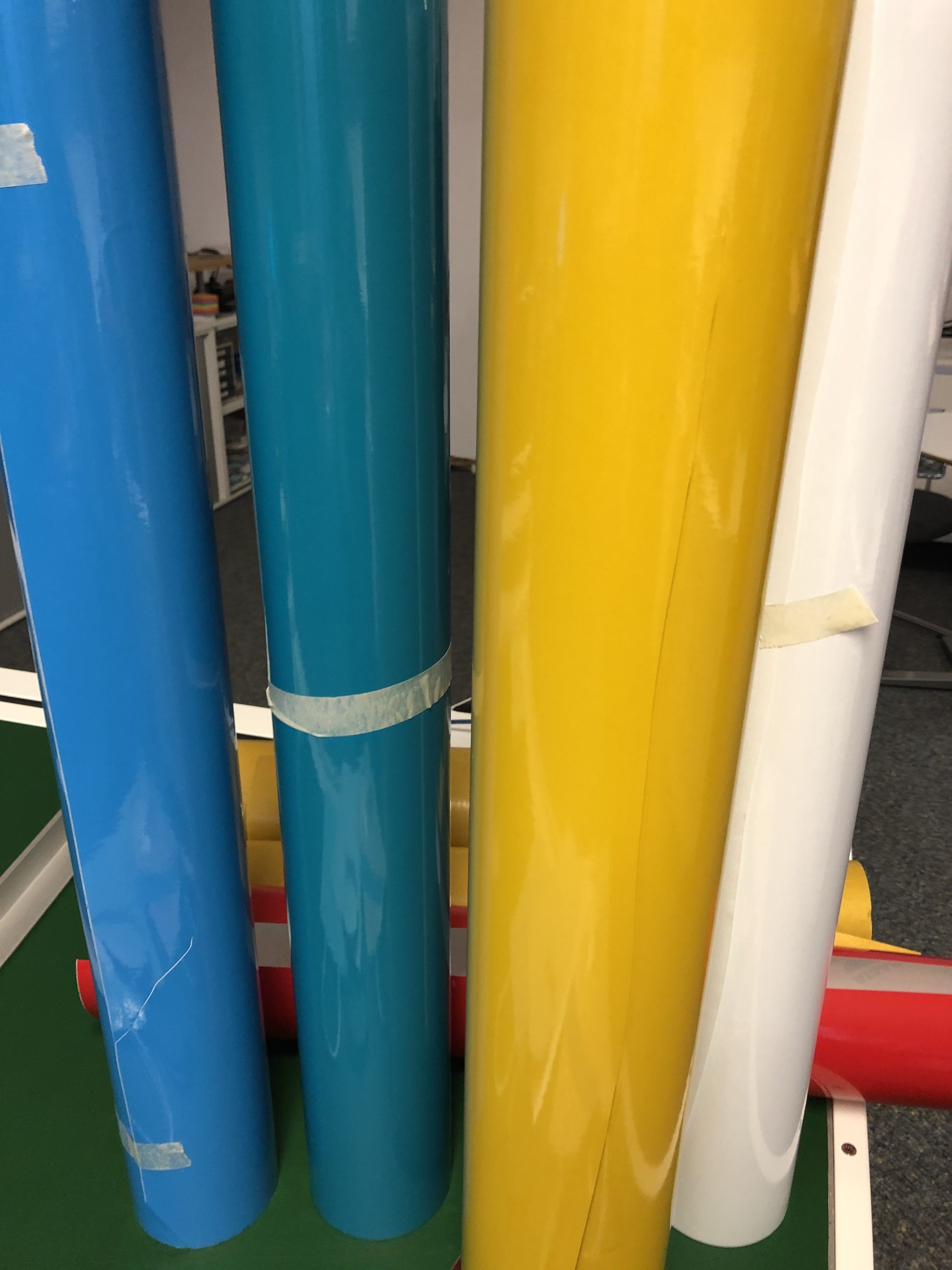 Aufrecht stehende Plotterfolienrollen in Hellblau, Petrol, Gelb und Weiß, dahinter liegend Rot – Farbauswahl für bunte Streifen auf der Tüte