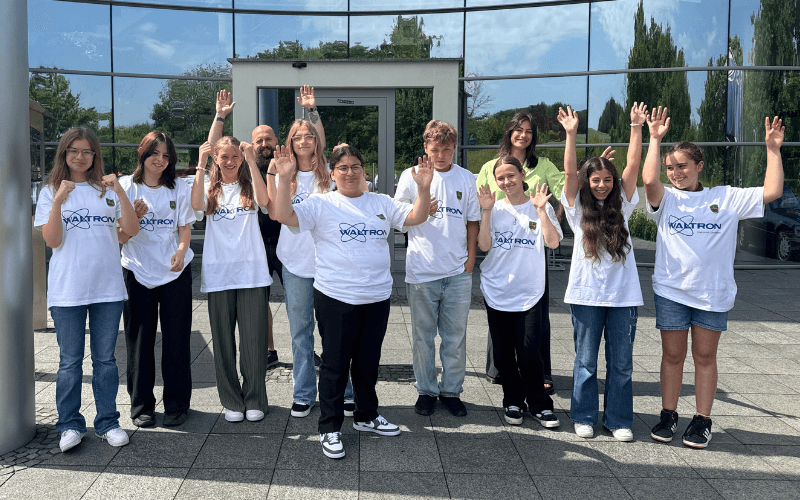 A group of pupils from Wolnzach Secondary School wave in front of a glass façade, wearing white T-shirts printed using DTF with the Waltron logo