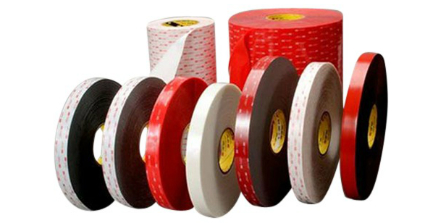 Rollos estrechos de cinta adhesiva de espuma de doble cara en negro, blanco y gris, algunos con lámina protectora roja; detrás, dos rollos madres.