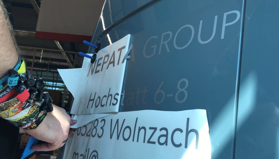 Hand zieht Übertragungsfolie vom grauen Transporter ab, darunter klebt Schriftzug NEPATA GROUP mit Adresse, blaue Magnete halten die Folie