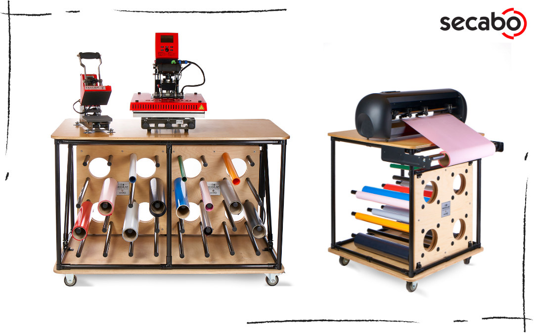 Zwei fahrbare Secabo Shelving Tables mit Folienrollen im Unterbau, oben zwei Transferpressen bzw. ein Schneideplotter mit rosa Folie