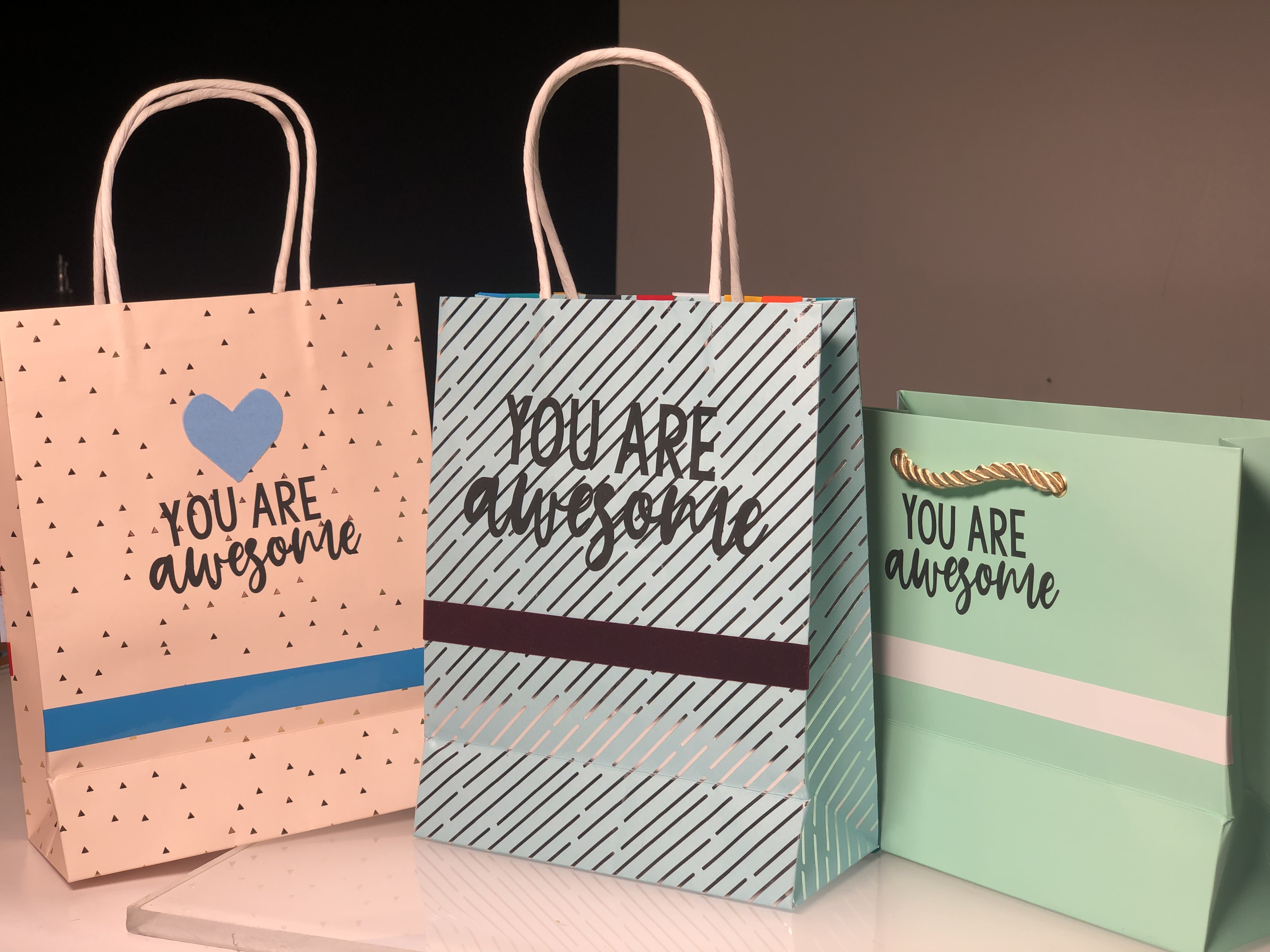 Drei Geschenktüten mit schwarzer Folienschrift You are awesome, verziert mit Folienstreifen in Blau, Bordeaux und Weiß sowie einem blauen Herz