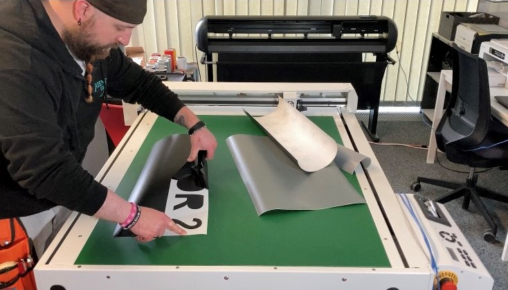 Mann entgittert schwarze Plotterfolie auf weißem Schild mit Schriftzug OR 2 am grünen Arbeitstisch, dahinter ein Schneideplotter