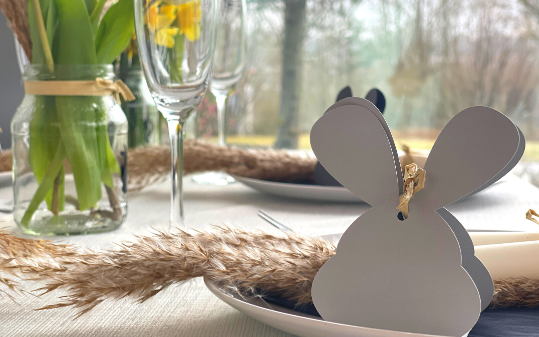 Geplottete Hasen-Geschenkverpackung aus hellem Papier mit Bastband auf Teller, Ostertisch mit Pampasgras, Tulpen im Glas und Sektgläsern