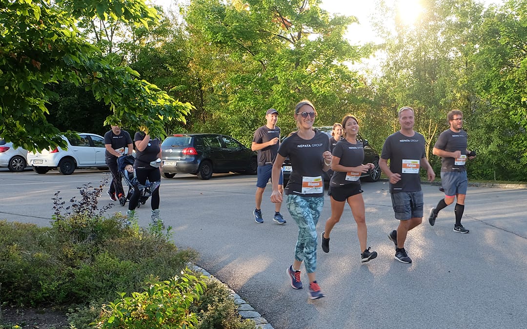 Nepata-Group-Team in schwarzen Shirts mit Startnummern beim virtuellen Firmenlauf im Abendlicht, hinten eine Teilnehmerin mit Kinderwagen