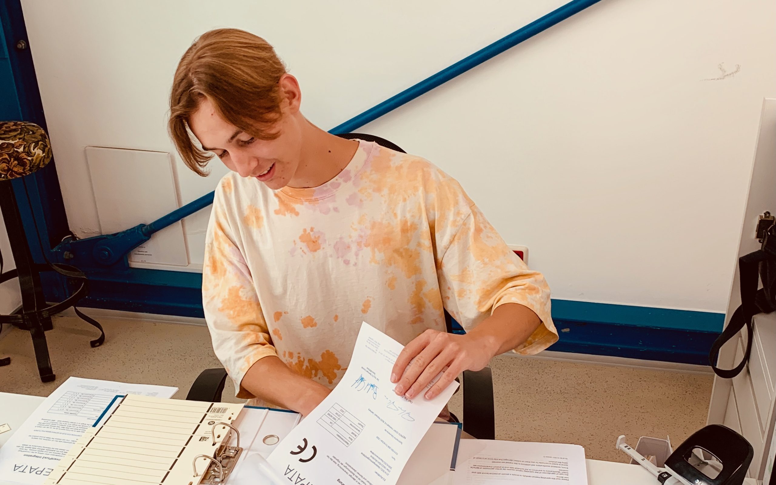 Praktikant Luca sortiert am Schreibtisch ein unterschriebenes CE-Dokument in einen Ordner mit Register ein, daneben ein Locher