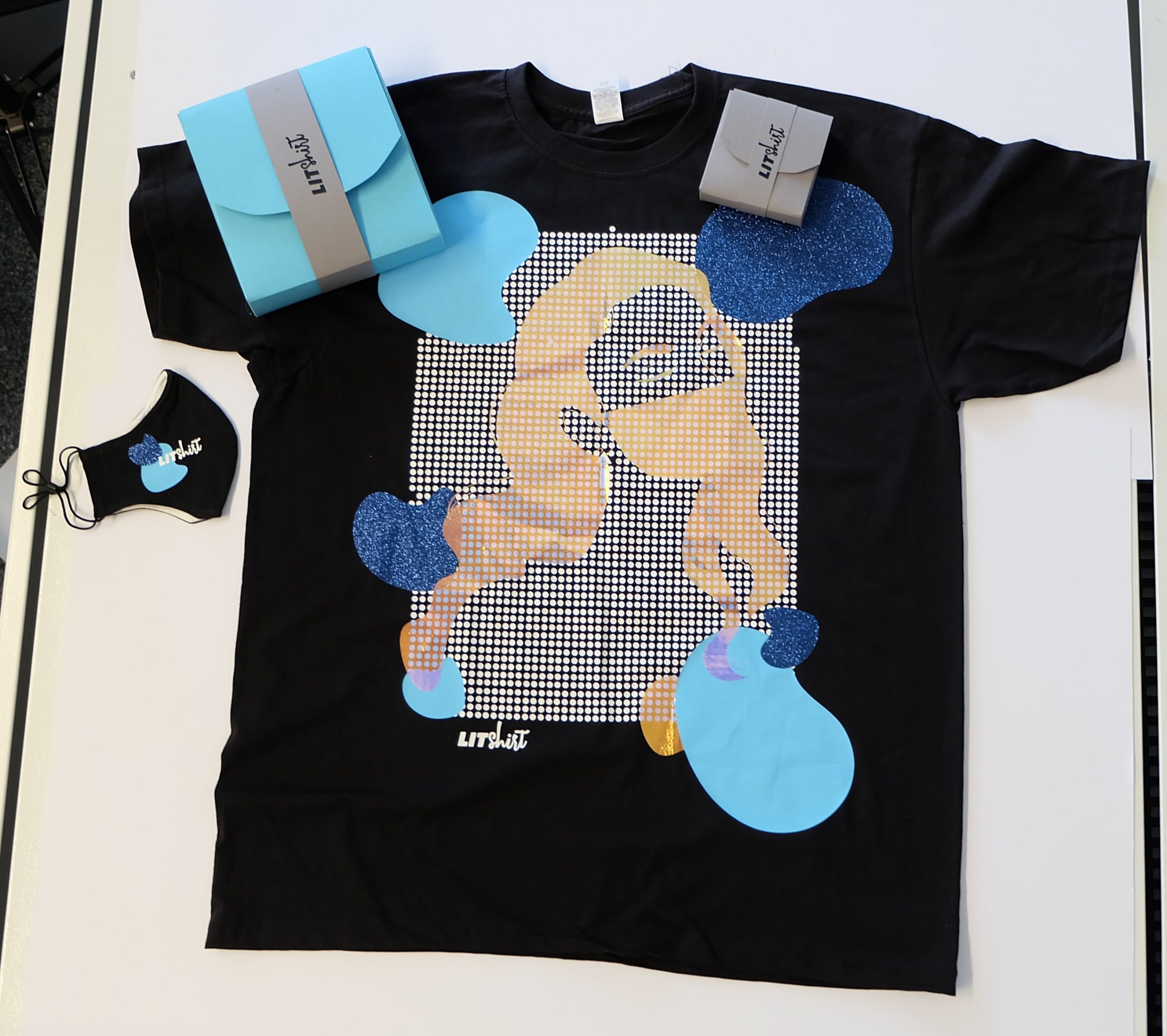 Schwarzes T-Shirt mit Folienmotiv aus Glitzer- und Metalliceffekten, dazu passende Maske und zwei geplottete LITshirt-Verpackungen in Blau und Grau