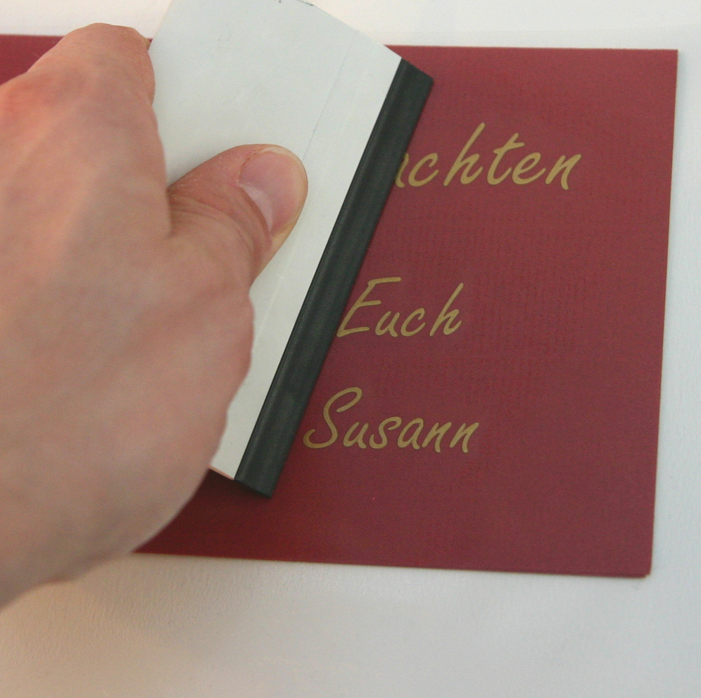 Hand drückt mit einer Rakel geplotteten Schriftzug in Gold auf eine rote Karte an, lesbar sind …achten, Euch und Susann