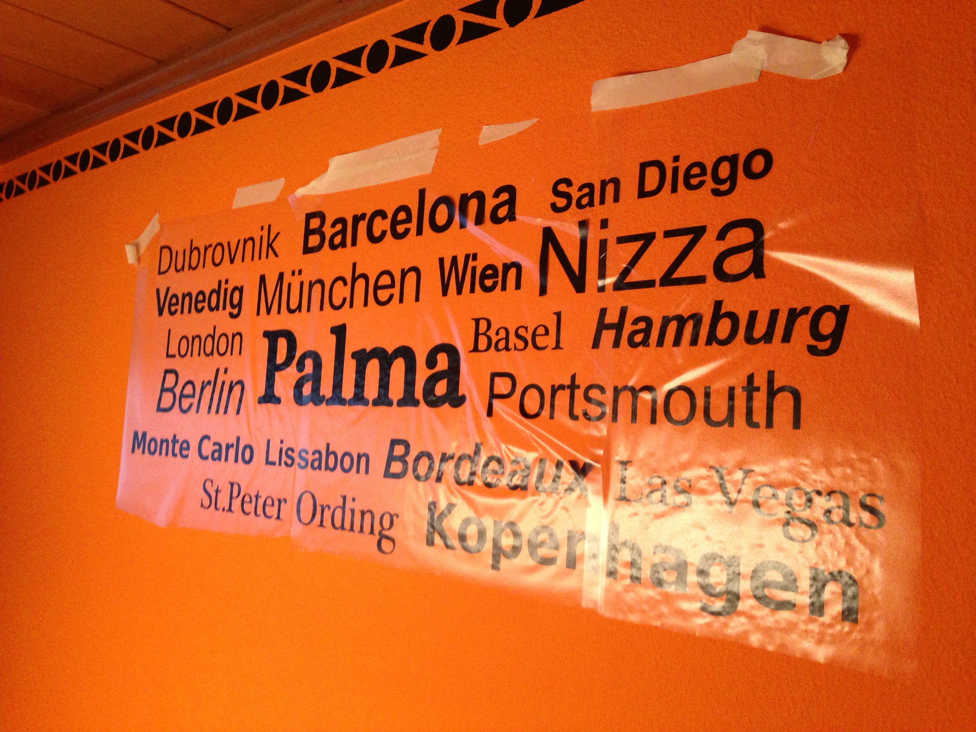Wandtattoo mit Städtenamen wie Palma und Nizza auf oranger Wand, noch mit transparentem Application Tape und Kreppband vor dem Abziehen