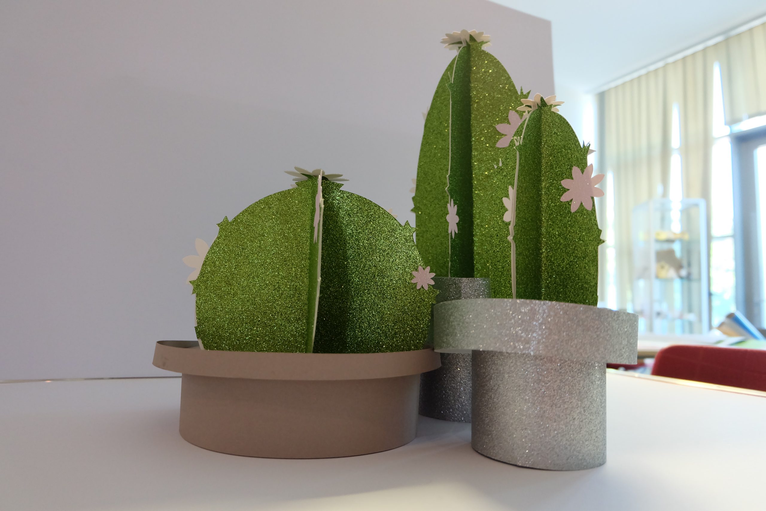 Drei 3D-Kakteen aus grünem Glitzerkarton mit weißen Blüten in Töpfen aus Karton und Silberglitzer, geschnitten mit dem Secabo FC100