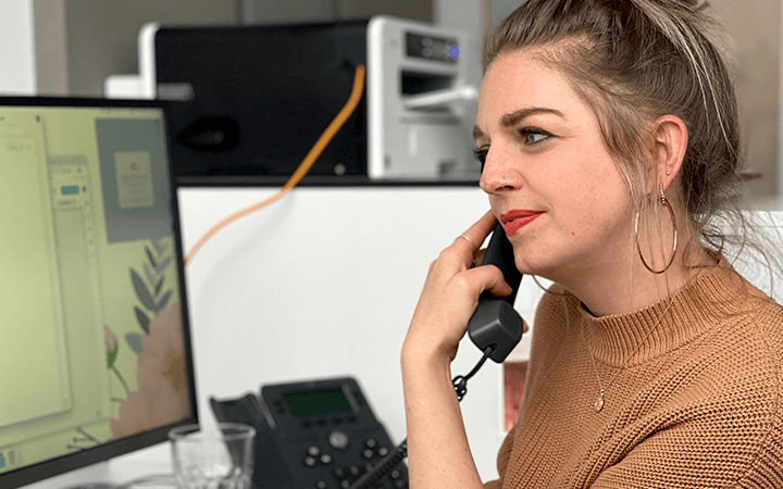 Neue Kollegin Sabrina im braunen Strickpullover telefoniert an ihrem Arbeitsplatz, daneben ein Monitor mit Grafikentwurf