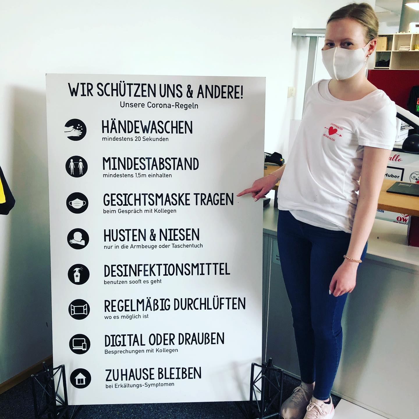 Frau mit Maske zeigt auf Tafel mit geplotteten Corona-Regeln: Wir schützen uns & andere! Händewaschen, Mindestabstand, Maske tragen u. a.