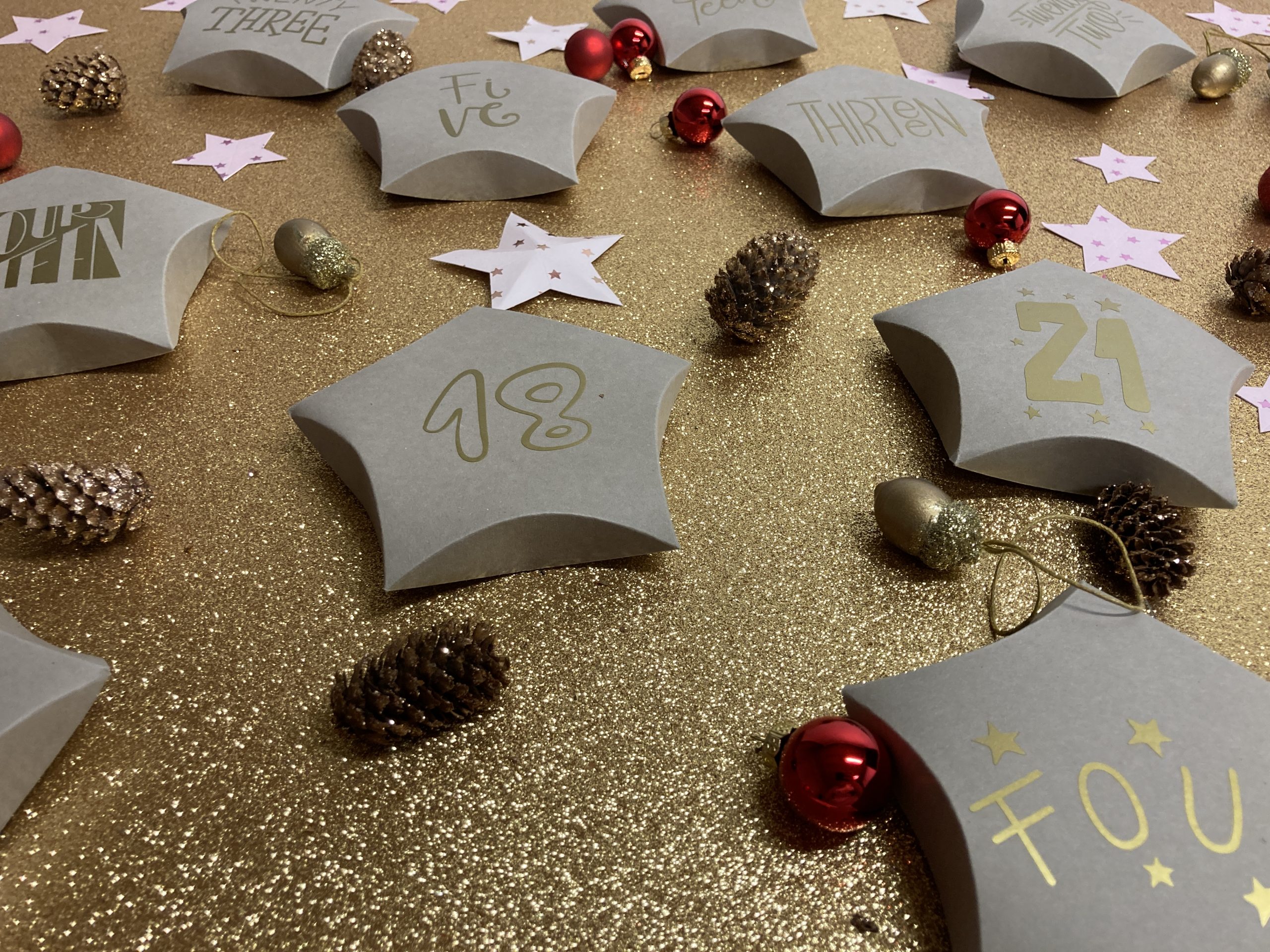 Graue Klapp-Sterne mit goldenen Zahlen wie 18, 21 und Five als Adventskalender, dazu Zapfen, rote Kugeln und rosa Papiersterne auf Goldglitzer