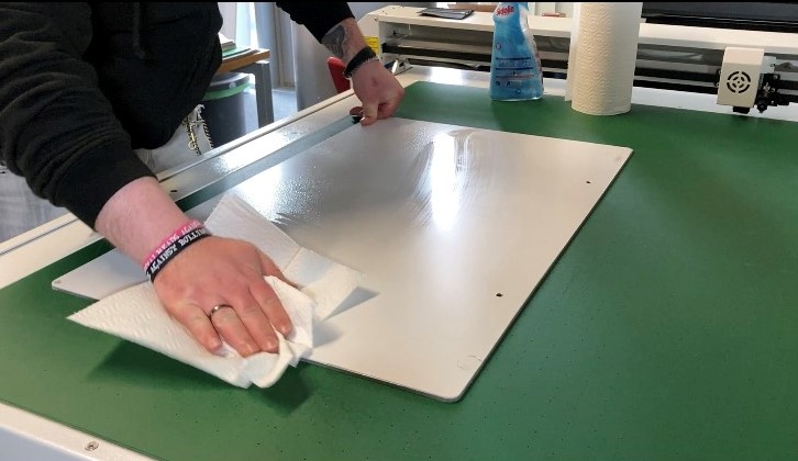 Hände reinigen ein weißes Schild mit Küchenpapier auf grüner Schneidematte, dahinter Glasreiniger und Küchenrolle – erster Schritt vor dem Kaschieren