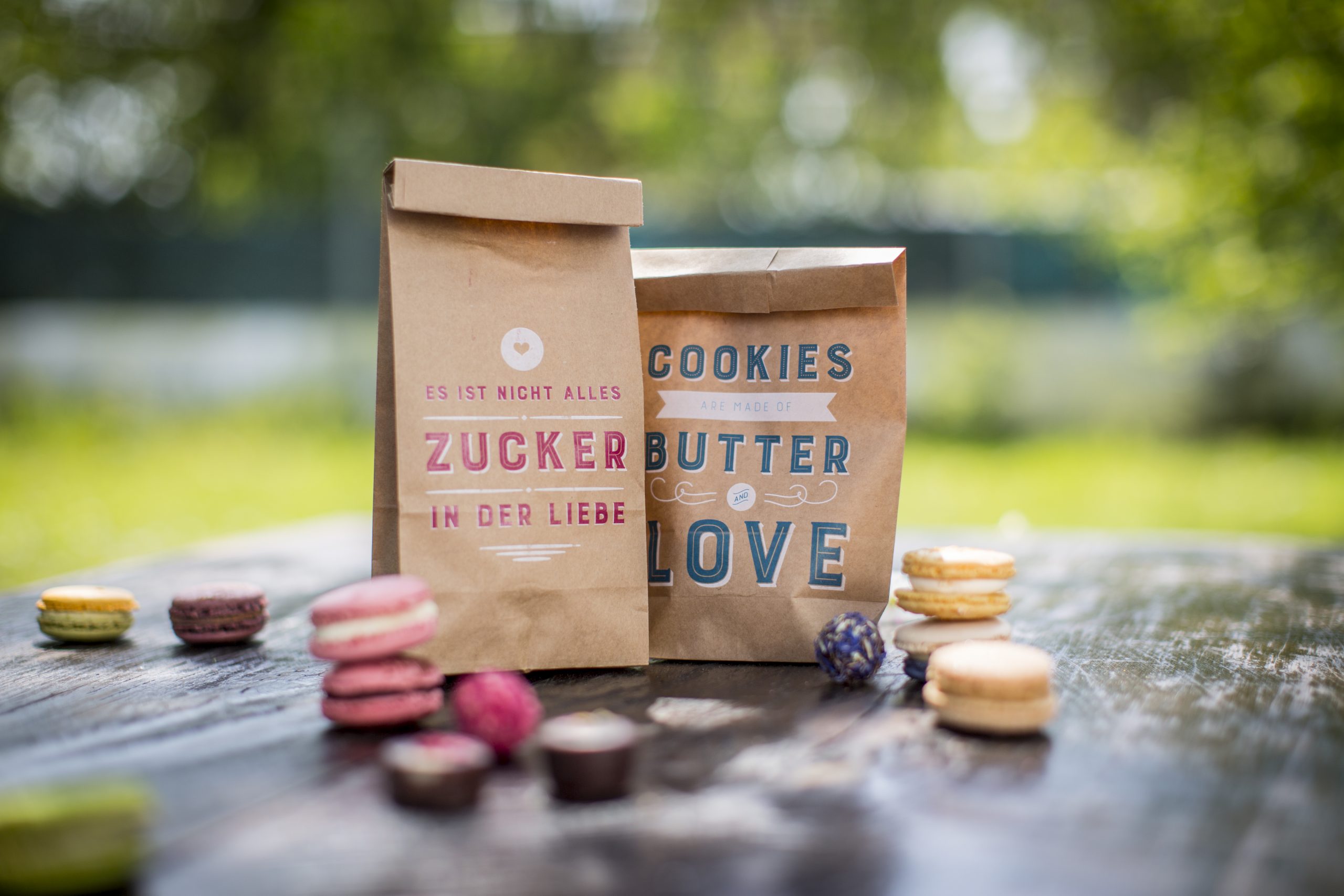 Zwei Kraftpapiertüten mit weiß unterlegtem Druck in Pink und Blau: Zucker in der Liebe und Cookies – Butter and Love, umgeben von Macarons