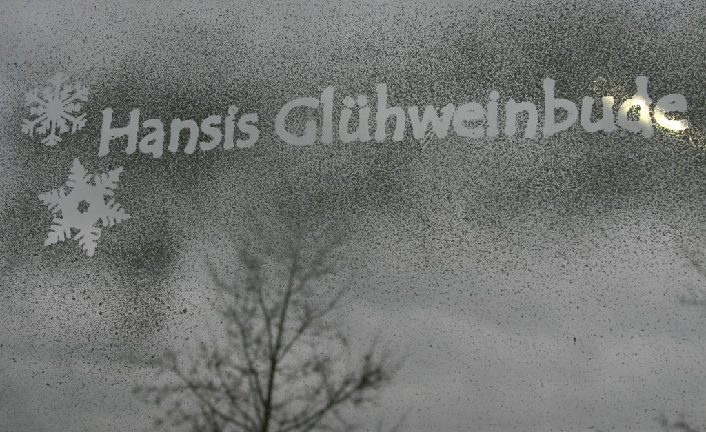 Fensterscheibe mit Snow-Spray, darin ausgespart der Schriftzug Hansis Glühweinbude und zwei Schneeflocken, gestaltet mit Maskierfolie