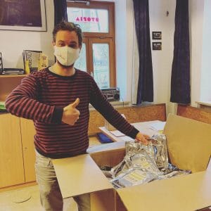 Mann mit Mund-Nasen-Schutz zeigt Daumen hoch neben einem Karton mit verpackten Masken, Teil der Spende von 2.000 James Masken an soziale Einrichtungen