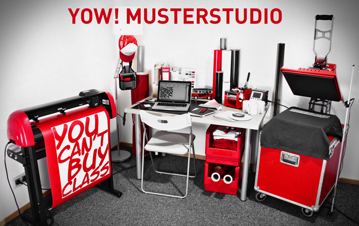 Beispielaufbau des YOW! Musterstudios: Schneideplotter, Schreibtisch mit Laptop, Tassenpresse, Transferpresse auf rotem Case und Folienrollen