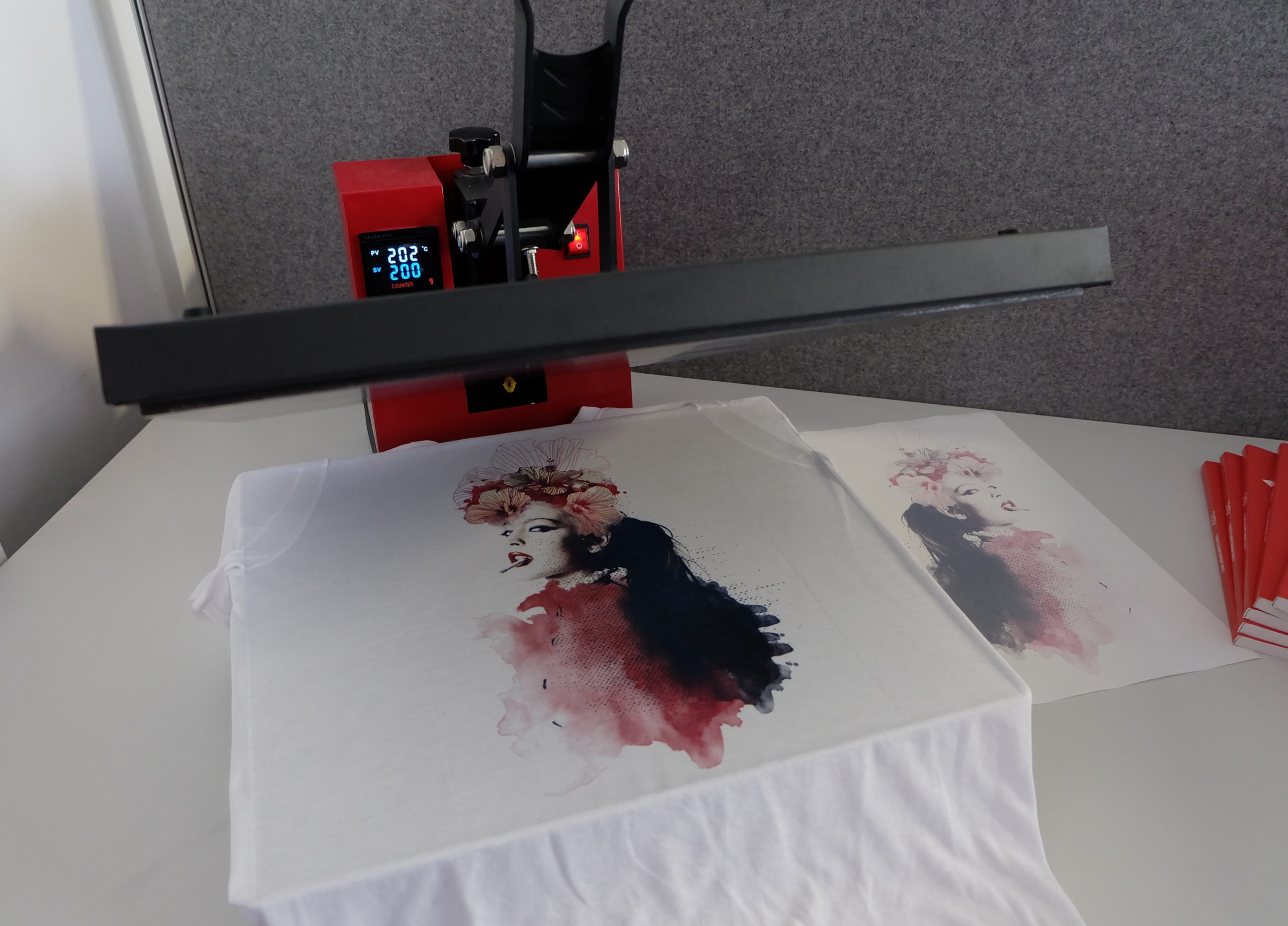 Transferpresse bei 200 °C über weißem T-Shirt mit sublimiertem Frauenporträt mit Blüten, daneben das verblasste Trägerpapier nach dem Transfer