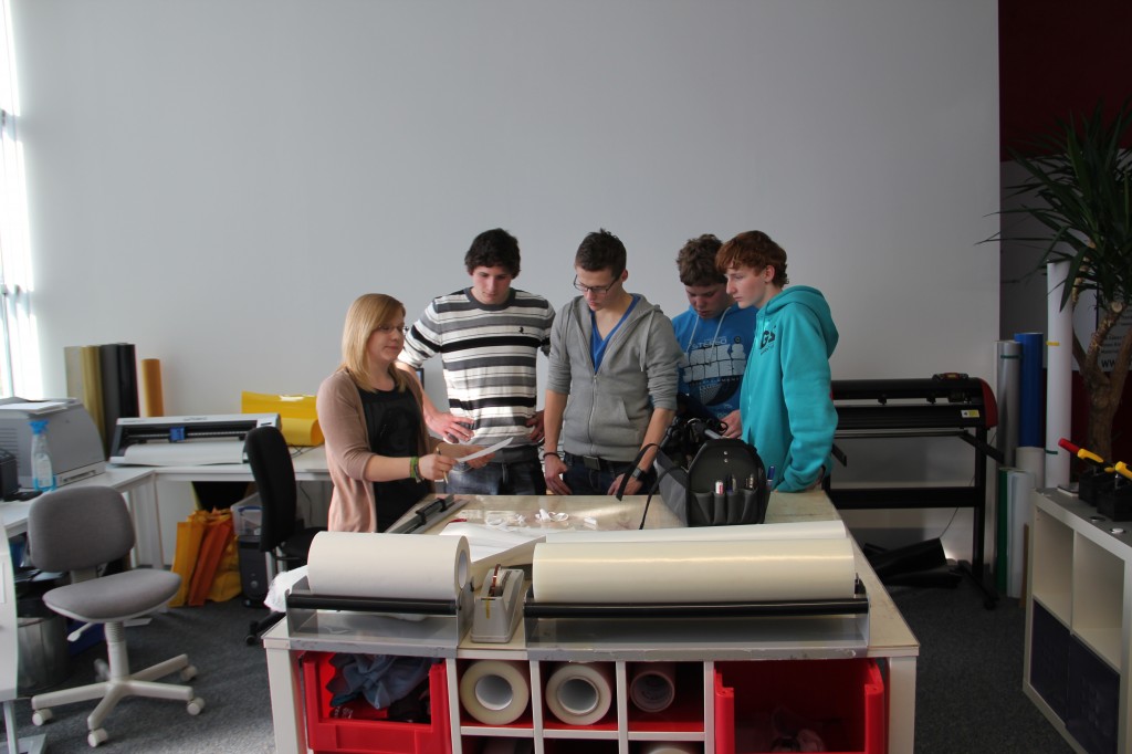 YOW!-Mitarbeiterin zeigt vier Schülern von YIP15 an einem Tisch mit Folienrollen die Folienverarbeitung, im Hintergrund ein Schneideplotter