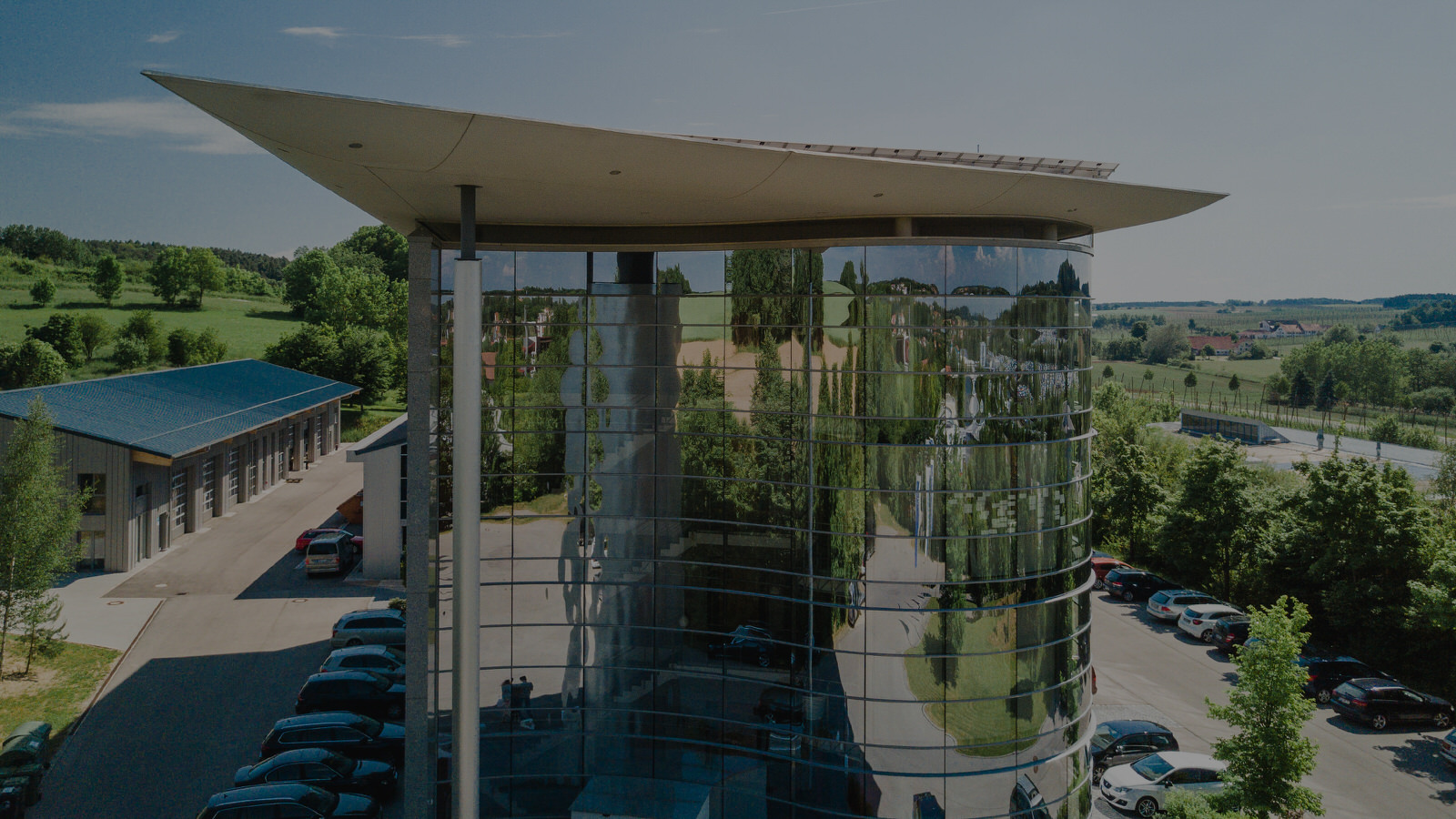 Bâtiment de la société NEPATA : construction ronde en verre réfléchissant, dotée d'un toit plat incurvé, flanquée d'un hall et de places de parking au milieu d'un paysage verdoyant