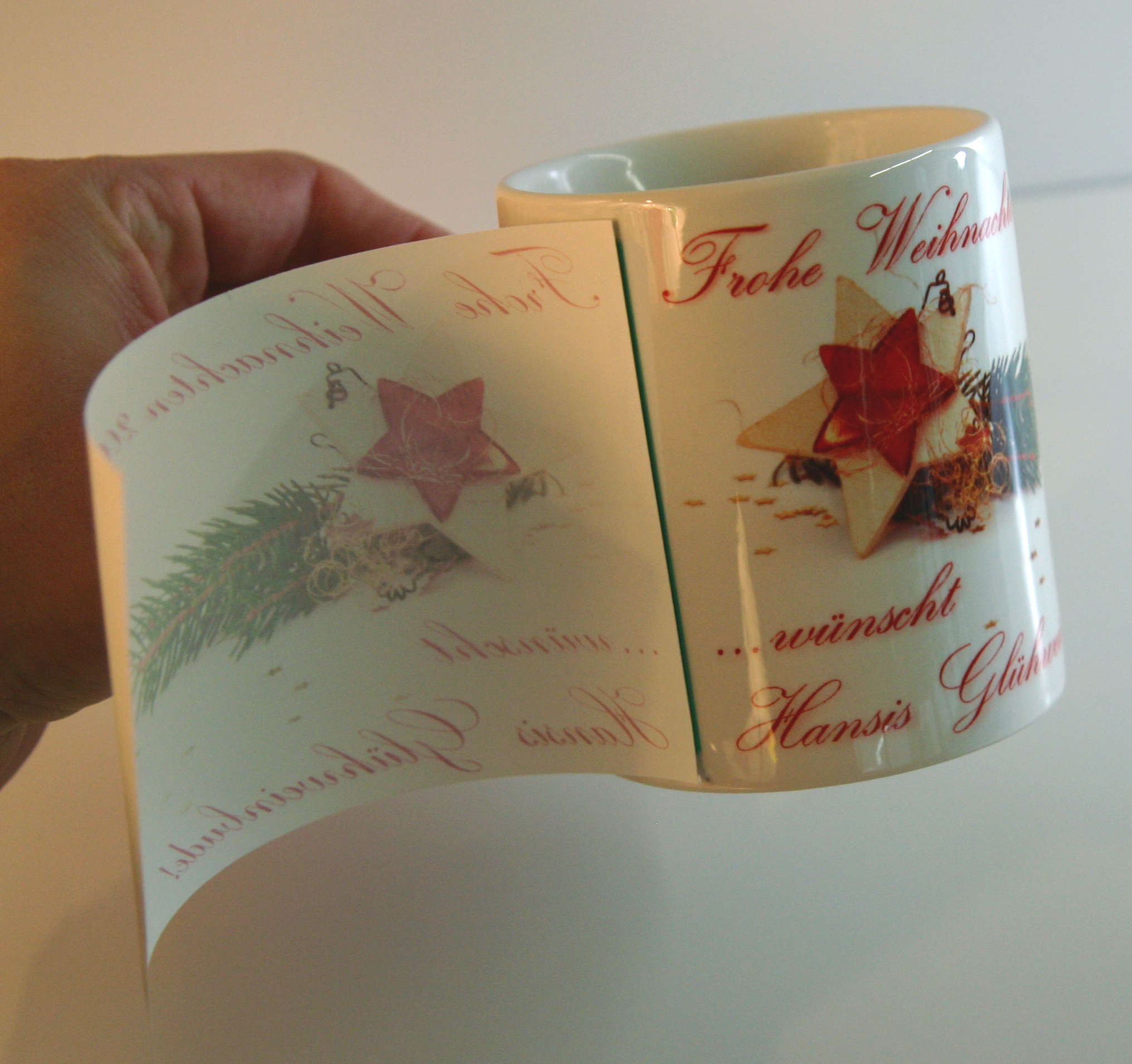 Hand zieht Sublimationspapier von Tasse ab, darunter Weihnachtsmotiv mit Sternen und Schriftzug Frohe Weihnachten … wünscht Hansis Glühwein