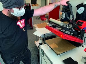 Mitarbeiter mit bedruckter Maske bedient eine rote Secabo-Transferpresse in der Werkstatt, um Masken oder Textilien zu bedrucken