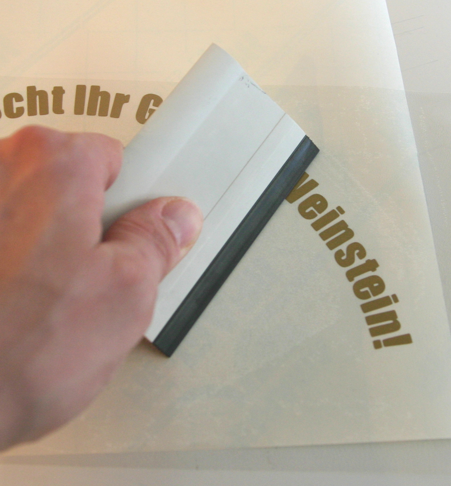 Hand streicht mit einem Rakel Übertragungsfolie auf den bogenförmig geplotteten goldenen Schriftzug für das weihnachtliche Geschirr