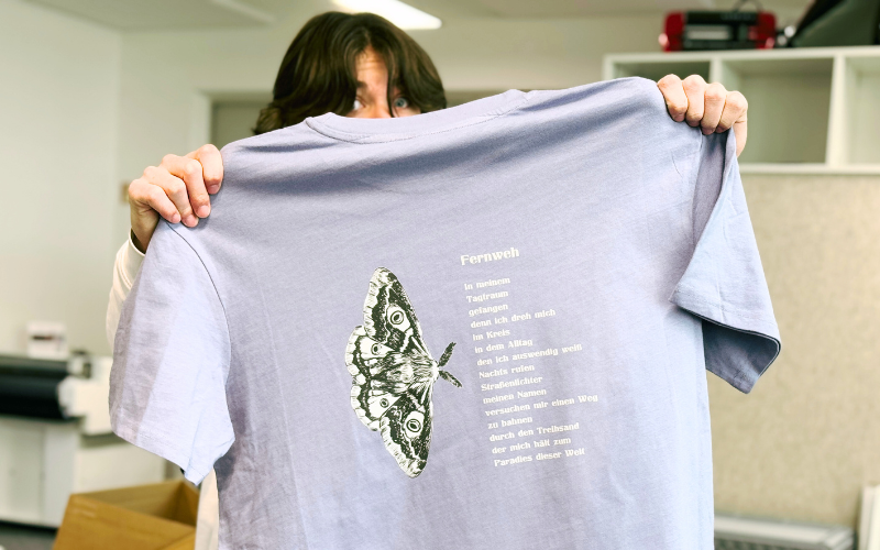 Person hält fliederfarbenes Midnight-Poets-Club-Shirt mit DTF-Druck hoch: Nachtfalter-Motiv und Songtext Fernweh in Weiß