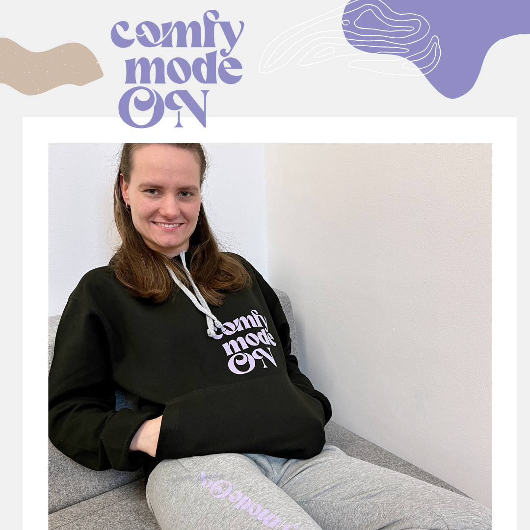 Frau auf Sofa in schwarzem Hoodie mit fliederfarbenem Flexdruck comfy mode ON und grauer Jogginghose mit Beinaufdruck als Textilveredelung
