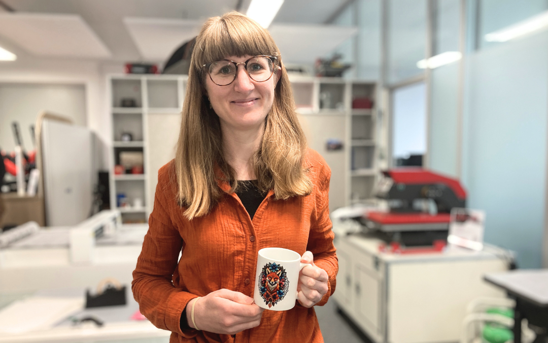 Kathrin in orangefarbener Bluse hält eine Tasse mit Fuchsmotiv im Nepata-Büro, im Hintergrund Regale und eine rote Transferpresse