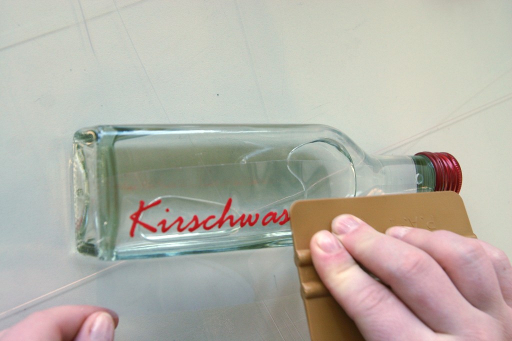 Hand rakelt den roten Flockfolien-Schriftzug Kirschwasser mit einer Rakel blasenfrei auf eine liegende Glasflasche mit rotem Schraubverschluss