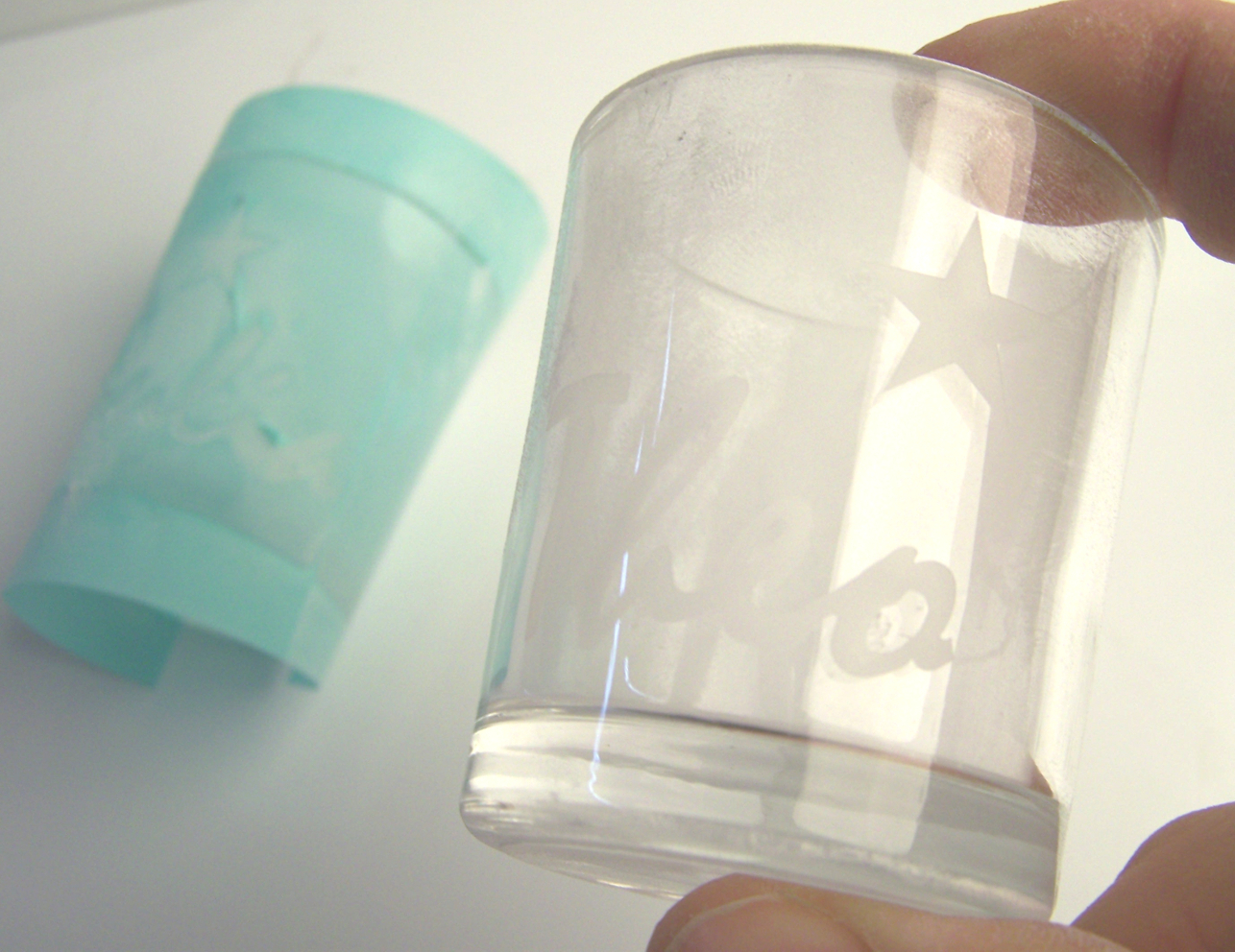 Teelichtglas mit sandgestrahltem Namensschriftzug und Stern in der Hand, dahinter die abgelöste türkisfarbene Maskierfolie