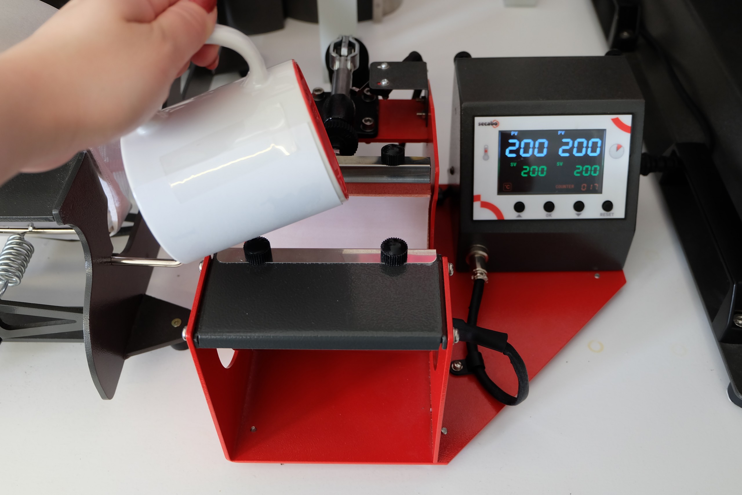 Weiße Tasse mit aufgeklebtem Transferpapier wird in die geöffnete Secabo TM1 Tassenpresse eingelegt, Display zeigt 200 °C