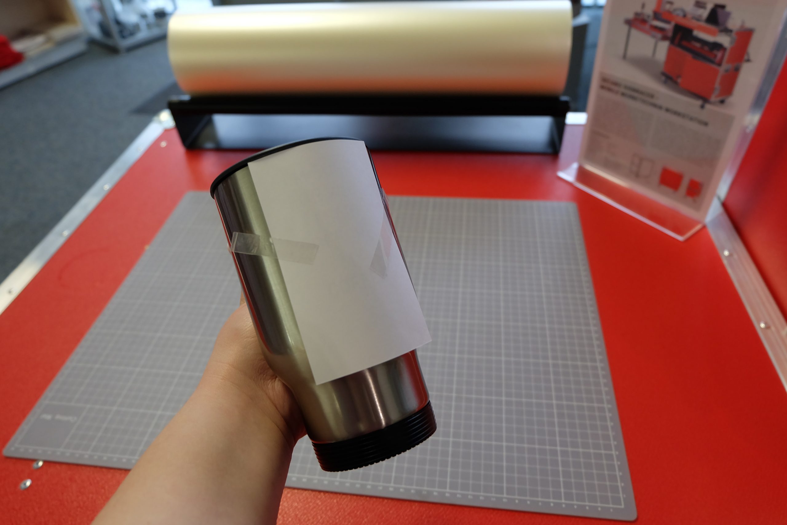 Hand hält Edelstahl-Thermobecher mit Sublimationspapier, das mit hitzebeständigem Klebeband fixiert ist, über einer Schneidematte