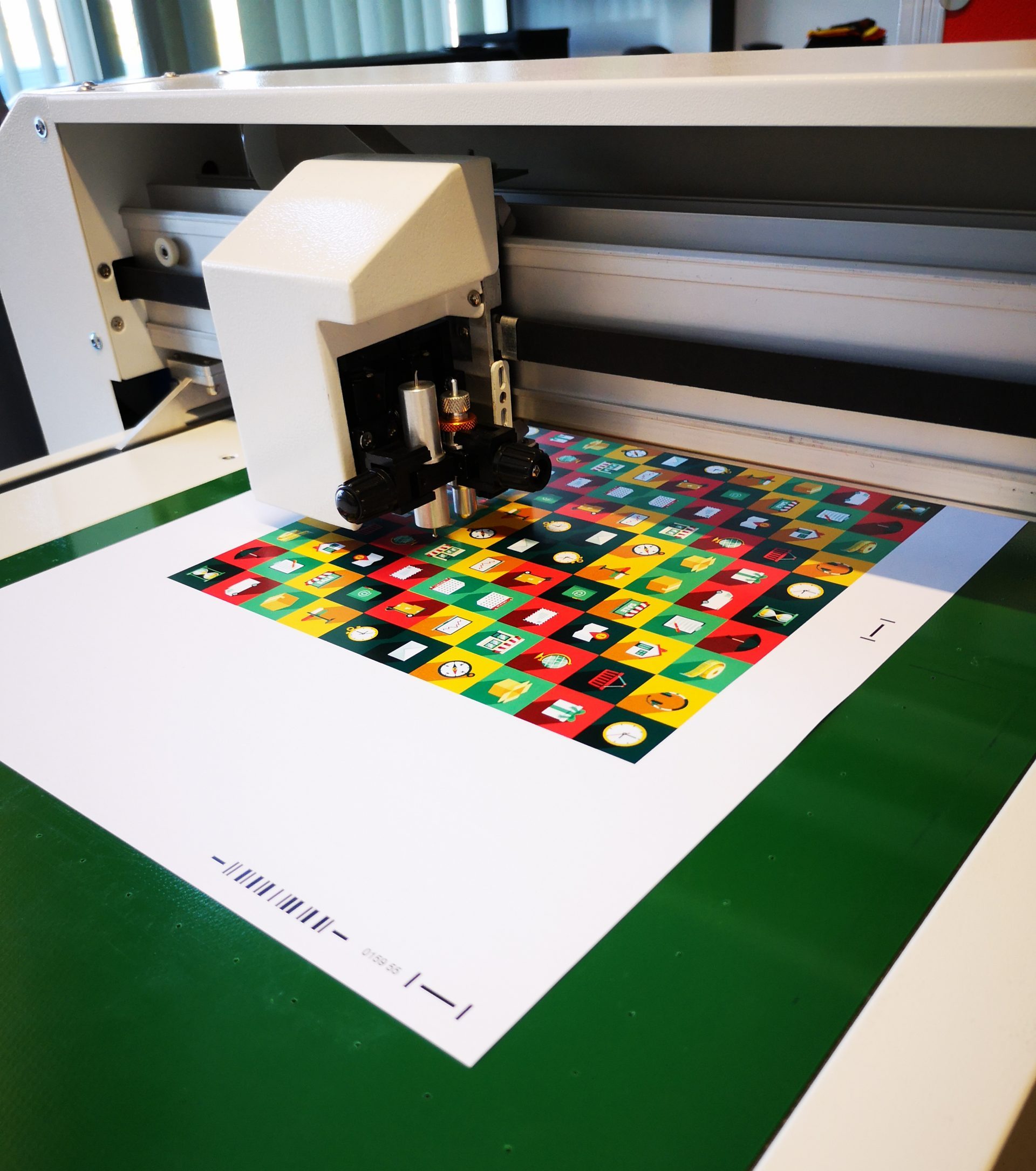 Schneideplotter FC 50 mit Messer- und Rillwerkzeug über bedrucktem Karton mit buntem Icon-Muster und Passermarken auf grüner Arbeitsfläche