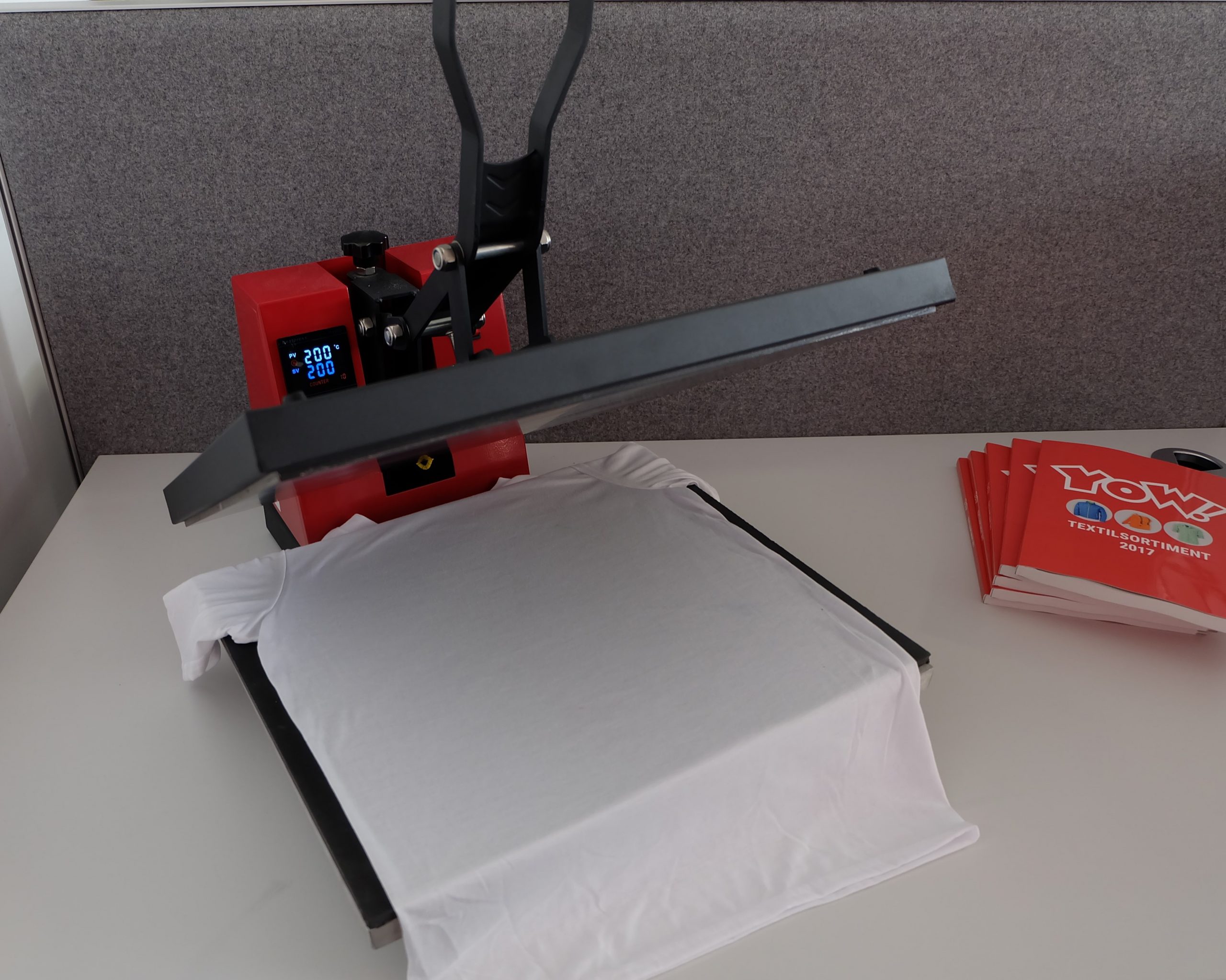 Geöffnete Transferpresse mit weißem Polyester-Shirt auf der Basisplatte, Display zeigt 200 °C Ist- und Solltemperatur, daneben YOW-Kataloge