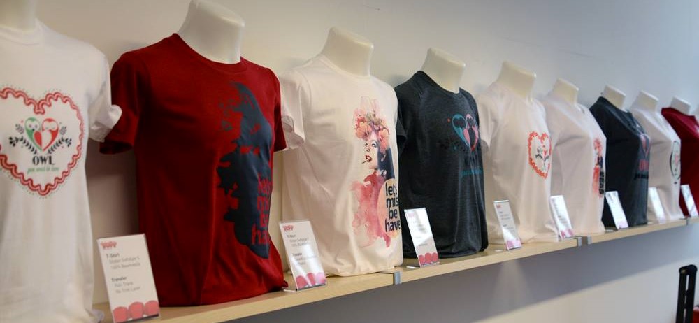 Bedruckte T-Shirts auf Büsten im Regal mit Infokärtchen, u. a. Eulen-Herzmotiv und Frauenporträt, als Beispiele für Textilveredelung mit Folie