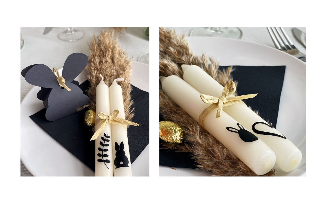 Zwei cremefarbene Stabkerzen mit schwarzen Folienmotiven wie Hase und Zweig, mit Bastschleife auf Teller mit Pampasgras, Goldei und Hasenpäckchen