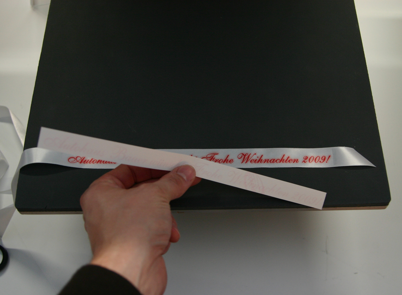 Hand zieht nach dem Pressen das Sublimationspapier vom weißen Geschenkband auf der Transferpresse ab – rot bedruckt: Frohe Weihnachten 2009!