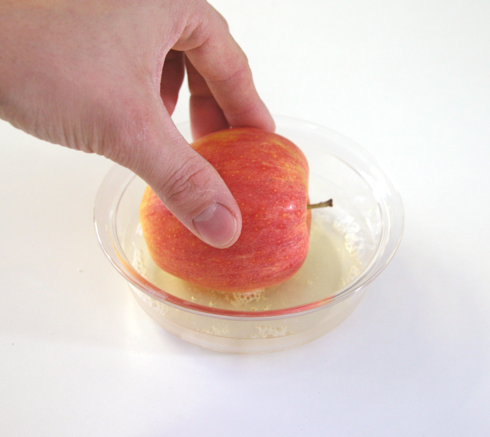 Hand taucht einen rot-gelben Apfel in eine Glasschale mit schaumigem Eiweiß – Schritt 2 der Anleitung für Winteräpfel mit Puderzucker