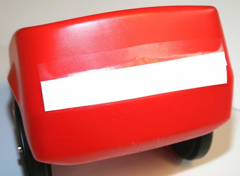 Rotes Bobby Car mit aufgelegtem Aufkleber samt Trägerpapier und Übertragungsfolie, beim Positionieren vor dem Festrakeln