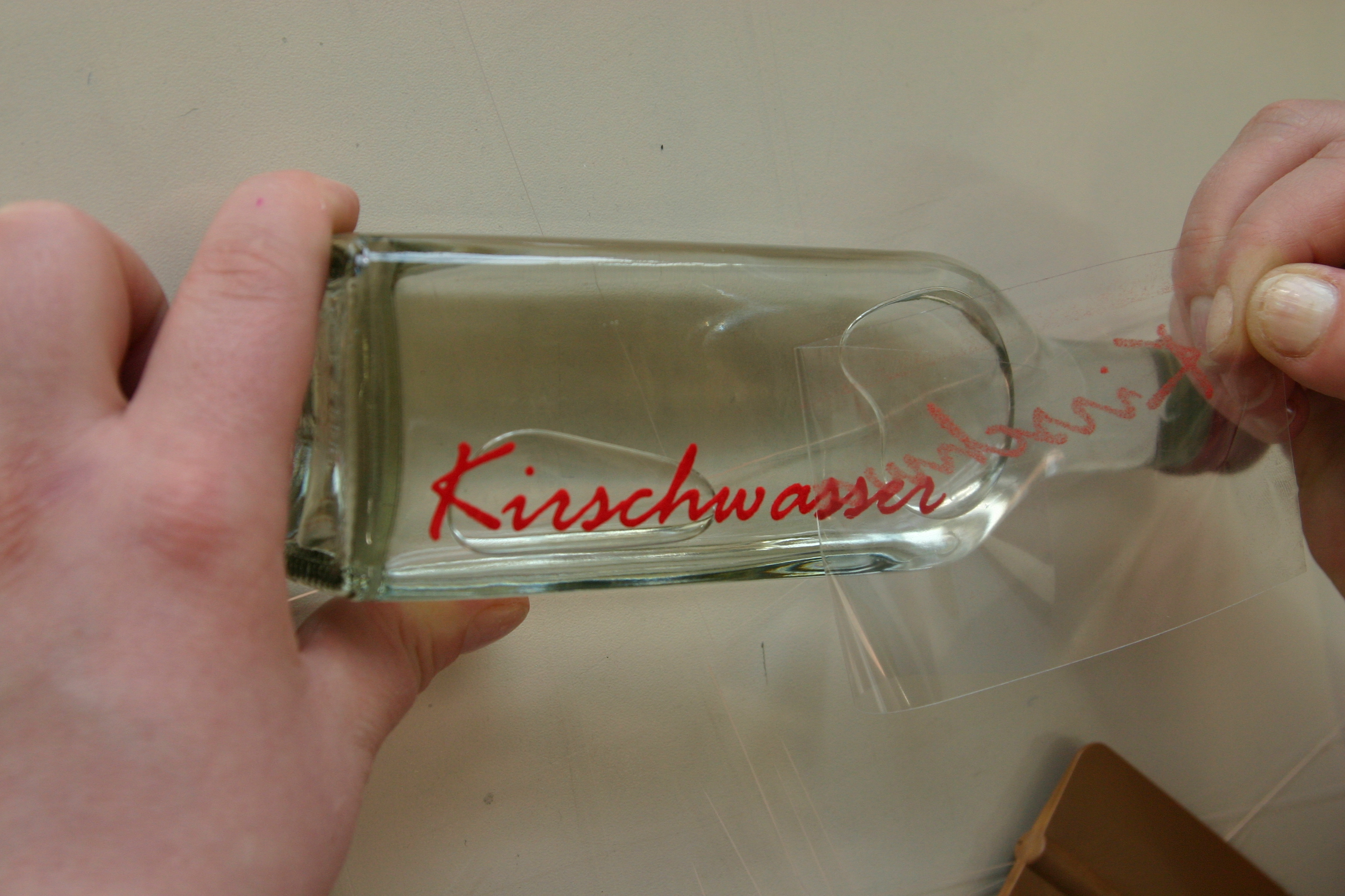 Übertragungsfolie wird von einer Glasflasche abgezogen, der rote Flockfolien-Schriftzug Kirschwasser bleibt auf dem Glas haften