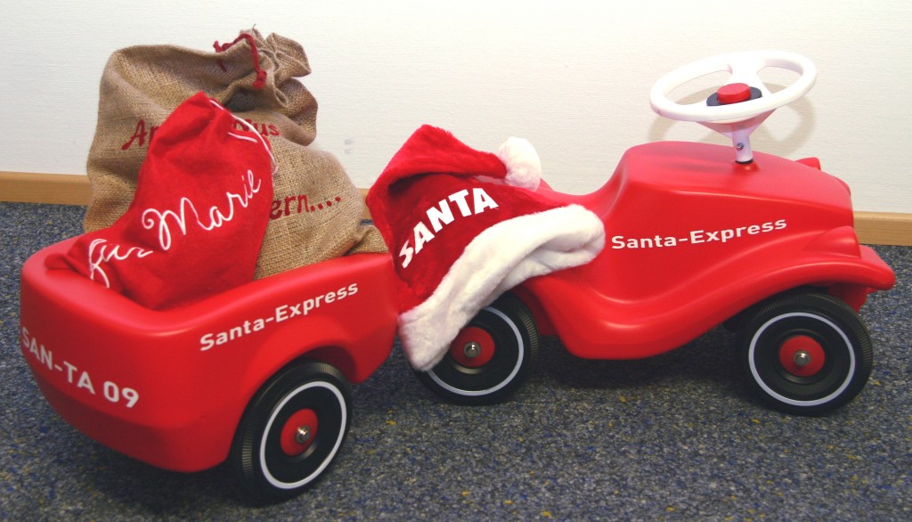 Rotes Bobby Car mit Anhänger, beklebt mit weißem Schriftzug Santa-Express und SAN-TA 09, dekoriert mit Nikolausmütze und Geschenksäcken
