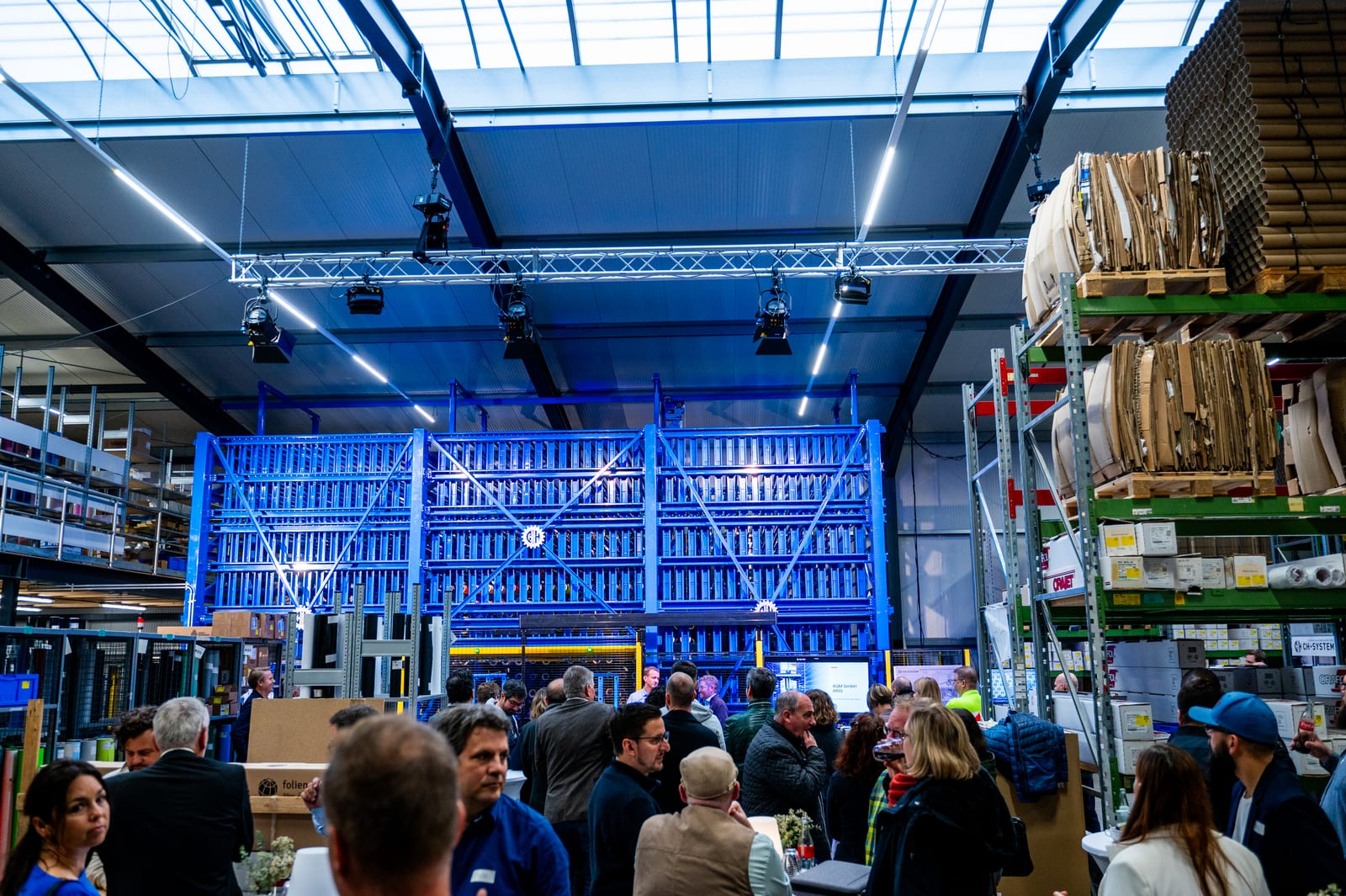 Premierengäste in der Lagerhalle von 4GM / Folienwelt in Trier vor dem blauen, mehrstöckigen Regal des automatischen Rollenlagers ARSS